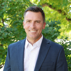 Man wearing a blazer in front of a tree smiling.